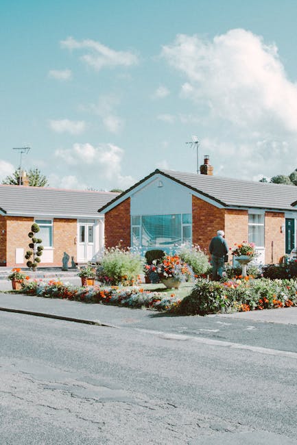 A single-story brick house with a grey tiled roof features a front garden with vibrant, well-maintained flower beds filled with colorful blooms and lush green shrubs. The garden layout includes a pave